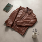 Brown leather jacket on a light gray surface with a notebook and small plant.